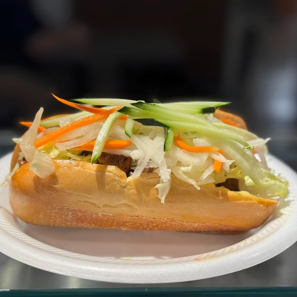 A banh mi sandwich on a white paper plate, filled with meat, lettuce, carrots, and cucumbers.