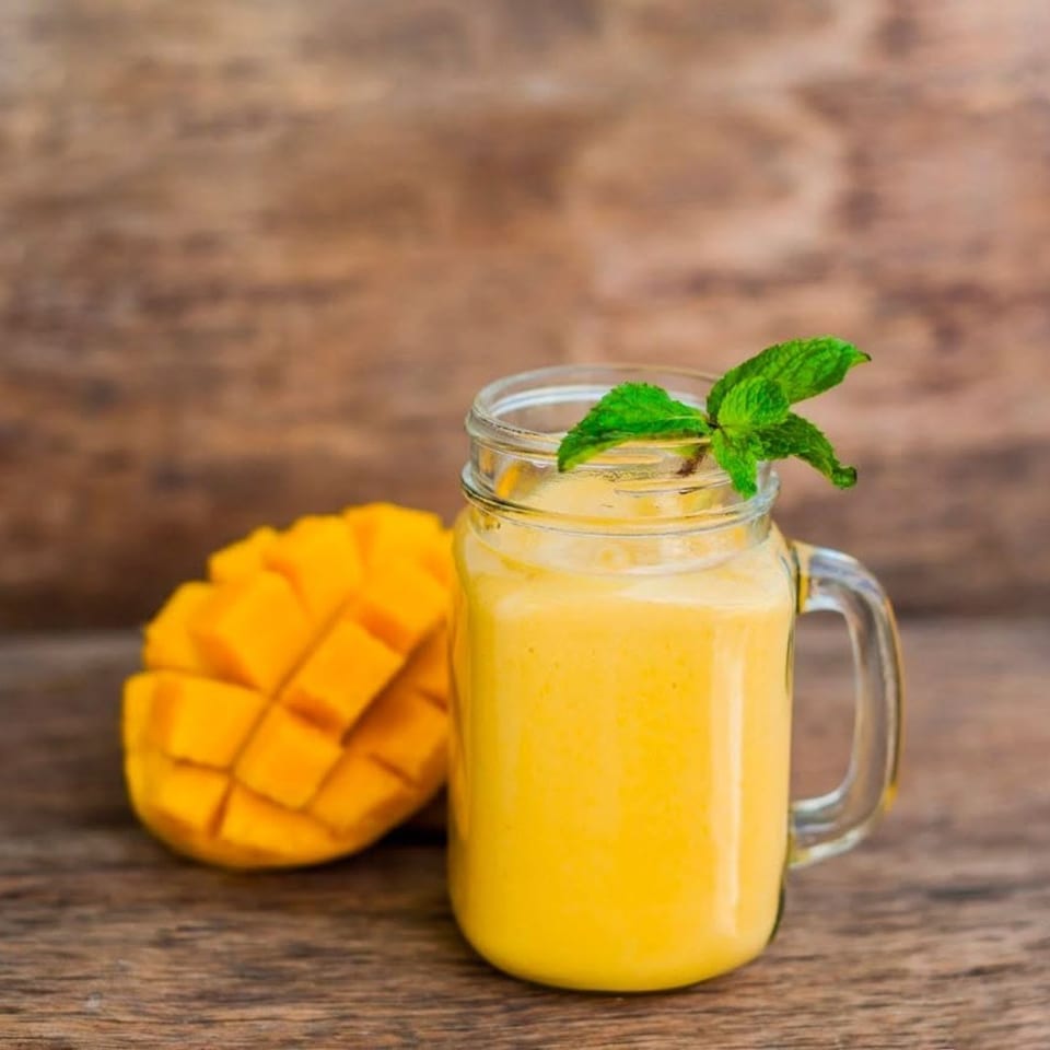 A mango smoothie in a mason jar with mint, next to a sliced mango on a wooden surface.