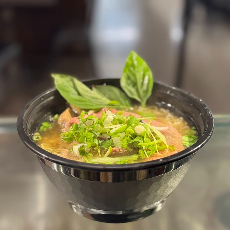 A black bowl of pho with beef, noodles, broth, green onions, and basil leaves.