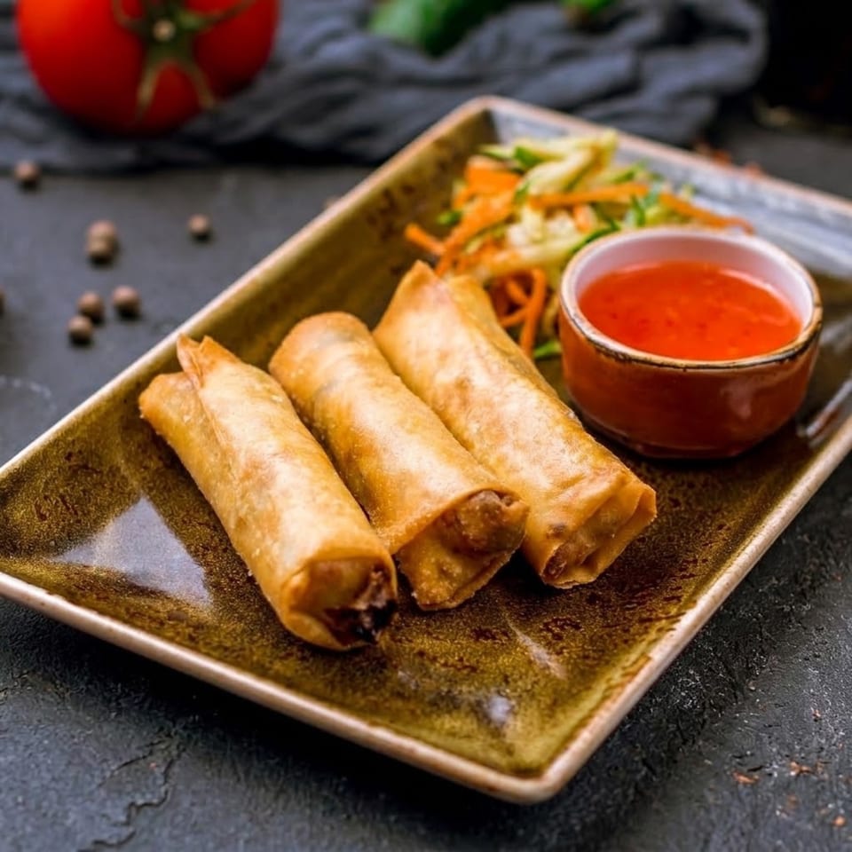 Three golden-brown spring rolls with a side of sweet chili sauce and shredded vegetables on a rectangular plate.